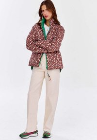 Brązowa pikowana kurtka w białe kropki, z zielonym zamkiem i kołnierzem; zestawiona z beżowymi spodniami i zielono-różowymi sneakersami; prosta koralikowa biżuteria.