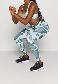 Un reggiseno sportivo blu chiaro e leggings con motivo mimetico nelle tonalità di blu e verde indossati durante uno squat. Scarpe da atletica nere con suole bianche.