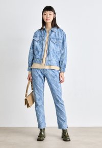 Jaqueta de denim azul clara com dois bolsos no peito, combinada com calças do mesmo tecido. Suéter bege por baixo e botas oliva. Bolsa de ombro camel.