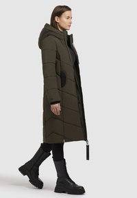 Olivově zelený prošívaný kabát s kapucí, s diagonálním prošíváním, černými akcenty a předním zipem. Spojen s černými šněrovacími botami.