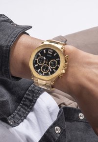 Auksinės nerūdijančio plieno laikrodis su juodu ciferblatu, aštuonkampiu korpusu, trimis subciferblatais ir dideliais numeriniais žymenimis. Turima nuorodų apyrankės dizainas.