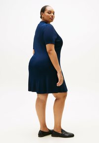 Marineblaues geripptes Kleid mit kurzen Ärmeln, tailliertem Oberteil und ausgestelltem Saum. getragen mit schwarzen Slippern. Einfarbiger Hintergrund.