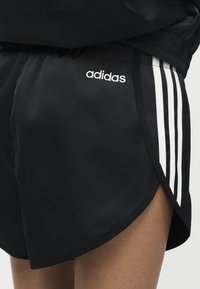 Μαύρο αθλητικό σορτς Adidas με λευκές τρεις ρίγες στο πλάι και λευκό λογότυπο Adidas κοντά στη μέση.