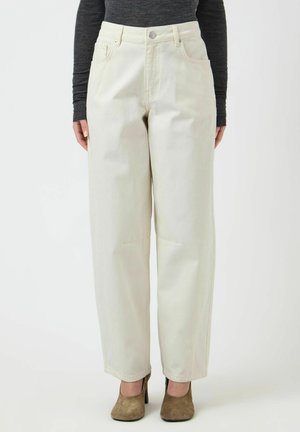 Kvinde iført cremefarvede bukser med højtaljet, vide ben og forlommer, kombineret med brune ruskindsstiletter og en mørk langærmet top.