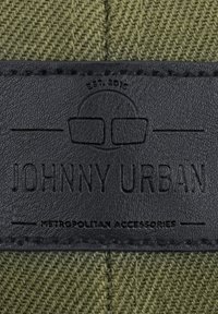 Fekete bőr címke, dombornyomott "JOHNNY URBAN" és "METROPOLITAN ACCESSORIES" felirattal, varrva textúrázott olajzöld anyagon, halszálka mintázattal.