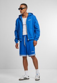 Blauer Kapuzenpullover mit Reißverschluss und Taschen; kombiniert mit blauen Shorts mit weißen Akzenten und Logo; gestylt mit Sonnenbrille.