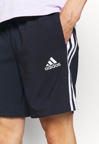 Persona que lleva pantalones cortos negros de Adidas con franjas blancas a los lados y el logotipo de Adidas, con la mano situada en el bolsillo derecho.
