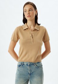 Polo shirt beige à manches courtes avec col, doté d'une patte de boutonnage et d'un ourlet côtelé, associé à un jean en denim bleu clair.