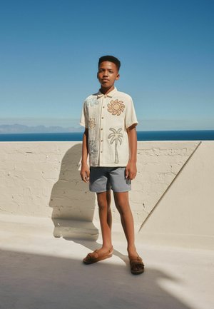 Dreng stående udendørs på en terrasse iført beige skjorte med sol- og palmedesign, grå shorts og brune sandaler under en klar blå himmel.