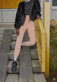 Černá umělá kožešinová bomber bunda, šedý rolák, růžové tepláky a černé tenisky s bílou podrážkou. Stojí na kamenných schodech vedle žlutého zábradlí.