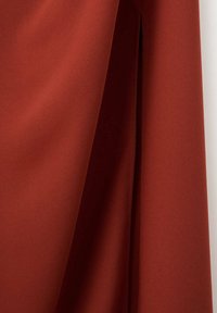 Rust-colored fabric with visible texture, hanging with soft folds and shadows against a white background.