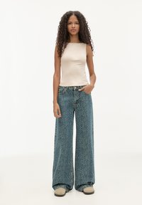 Lys beige ærmeløs top, parret med brede denim jeans med et struktureret blåt mønster. Model poserer med den ene hånd i lommen.