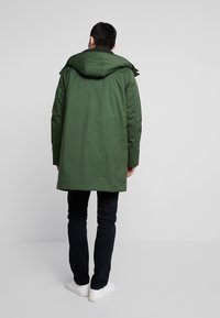 Abrigo parka verde con capucha, mangas largas y corte recto. Presenta una textura suave y un diseño sencillo. Se lleva con jeans oscuros y zapatillas blancas.