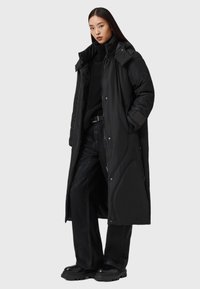 Czarna ocieplana kurtka z wysokim kołnierzem i zapięciem na zamek. Posiada gładką strukturę, boczne kieszenie oraz długi, luźny krój. Noszona na czarny strój.