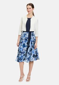 Marineblauwe jurk met een witte cropped jas. Rok met een blauw bloemenpatroon, tot de knie. Model draagt witte sleehakken.