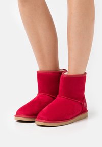 Bottines rouges en daim avec un bout arrondi, comportant des accents cousus et une semelle en caoutchouc beige. Doublure douce visible au bord supérieur.