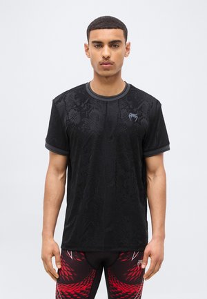 Άνδρας με κοντά μαλλιά, που φοράει μαύρο t-shirt με σχέδιο από δέρμα φιδιού και κολάν με κόκκινο-μαύρο μοτίβο, μπροστά σε απλό ανοιχτόχρωμο φόντο.