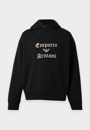 Νέος άνδρας που φοράει μαύρο φούτερ Emporio Armani, ανοιχτό μπλε τζιν και λευκά αθλητικά παπούτσια, στέκεται με το ένα χέρι στην τσέπη.