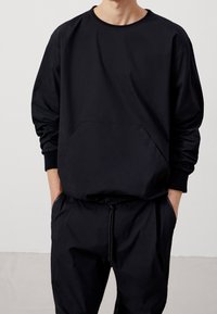 Felpa nera con maniche lunghe, scollo rotondo e tasca frontale. Abbinata a pantaloni neri coordinati con vita regolabile con coulisse.