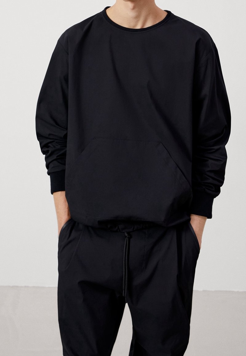 Felpa nera con maniche lunghe, scollo rotondo e tasca frontale. Abbinata a pantaloni neri coordinati con vita regolabile con coulisse.