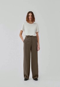 Woman with short brown hair wearing a white short-sleeve top and high-waisted wide-leg brown trousers standing with one hand in pocket.