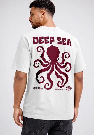 Man wearing white t-shirt with large maroon octopus and text "DEEP SEA" on the back, standing against a plain background.