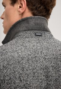 Cappotto di lana grigio a spina di pesce con un morbido colletto grigio. Presenta un'etichetta nera cucita sul retro sotto il colletto.