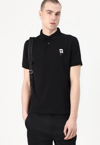 Fekete pamut póló, három gombos gallérral és egy kis fehér grafikával a bal mellrészen. Slim fit fazon.