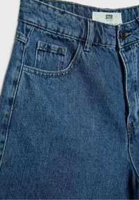 Blauer Jeansstoff im Nahaufnahme, zeigt die Vordertasche, Gürtelschlaufen, Knopfverschluss und das STR Teen Etikett im Bundinneren.