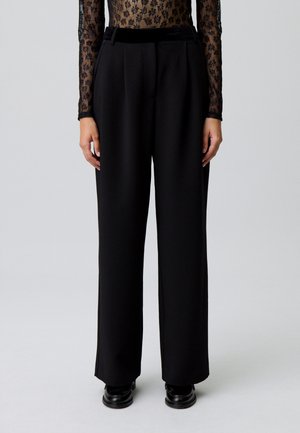 Persona vestida con pantalones anchos de cintura alta negros, una blusa de mangas largas de flor transparente metida por dentro y zapatos negros, de pie sobre un fondo neutro.