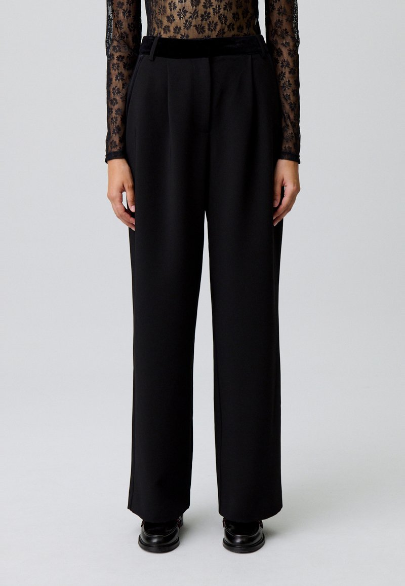 Personne portant un pantalon ample taille haute noir, un haut floral à manches longues transparent rentré à l'intérieur, et des chaussures noires se tenant sur un fond neutre.