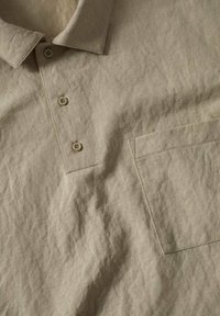 Polo en lin beige avec trois boutons, un col et une poche poitrine sur un tissu froissé.