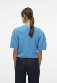 Maglione lavorato a maglia blu chiaro, a maniche corte, con una texture morbida, orlo a coste e scollatura arrotondata, indossato sopra jeans in denim scuro.
