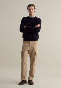 Maglione a girocollo lavorato a maglia blu navy, pantaloni cargo beige con tasche laterali, stivaletti marroni alla caviglia, modello in piedi con le braccia incrociate su uno sfondo neutro.