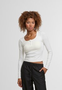 Witte lange mouwen crop top van ribbelstof met een ronde halslijn en drievoudige knoopsluiting; gecombineerd met zwarte broek met trekkoord.