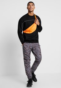 Melns kapuces krekls ar baltām niansēm, pelēki kamuflāžas bikses ar brūnām rakstām, un oranža plecu soma. Melni sporta apavi noslēdz izskatu.