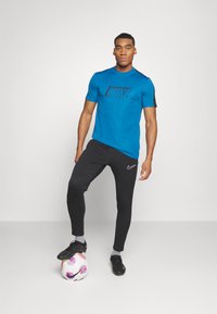 Camiseta corta de manga azul de Nike con logotipo negro, combinada con pantalones deportivos negros que tienen un detalle blanco de Nike, zapatillas negras y un balón de fútbol.