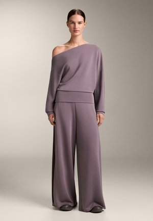 Femeie în picioare, purtând pantaloni largi mauve și largi și un top cu mâneci lungi, decoltat pe umeri, pe un fundal bej simplu.