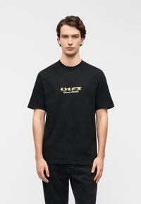 Μαύρο βαμβακερό μπλουζάκι με κοντά μανίκια, με λευκό και χρυσό γράφημα "LYLE'S Corner Market" στο κέντρο του στήθους.