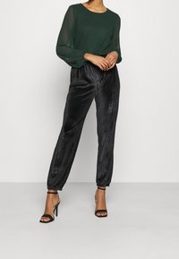 Grüne Bluse mit langen Ärmeln, kombiniert mit schwarzen Jogginghosen aus Samt. Die Bluse hat transparente Ärmel; die Jogginghosen verfügen über elastische Bündchen und eine glatte Textur.