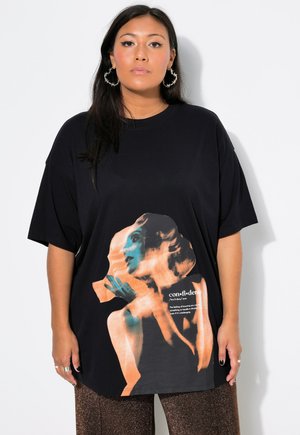 Mujer con cabello largo y oscuro lleva grandes pendientes de aro y una camiseta negra de gran tamaño con una imagen artística de una mujer y la palabra confianza.
