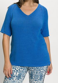 Modrý krátký rukáv svetr s výstřihem do V, s hladkým povrchem. V kombinaci s vzorovanými kalhotami s modro-bílými květinovými motivy.