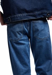 Jeans de denim en azul medio con un diseño clásico de cinco bolsillos. Presenta un ligero desgaste y una superficie texturizada; los bolsillos están cosidos.