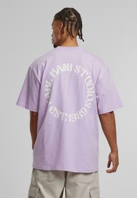 Lilac oversized t-shirt with white circular text "KANI STUDIOS EST. 1989" on the back. Fabric appears soft with a relaxed fit.