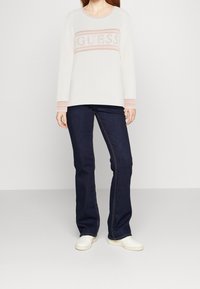 Haut blanc à manches longues avec "GUESS" en rose, associé à un jean foncé évasé et des baskets blanches. Tenue simple et décontractée.