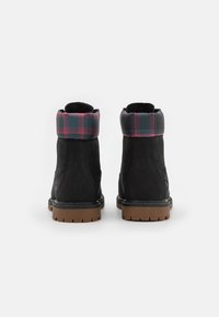 Botas de tobillo de ante negro con un puño de estampado a cuadros en tonos rosa y verde, suela marrón resistente con un diseño con relieve, vista trasera.