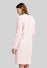 Mujer vestida con un vestido largo de manga larga rosa claro con un ribete de encaje blanco en el cuello y los puños, de espaldas contra un fondo gris.