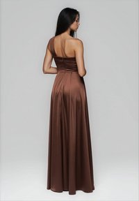 Donna con lunghi capelli scuri che indossa un abito in satin marrone, fino al pavimento, a spalla unica, vista di spalle contro uno sfondo grigio chiaro uniforme.