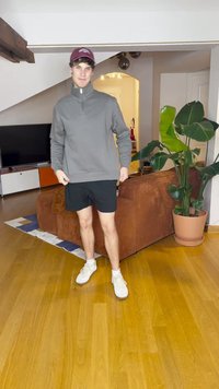 Felpa grigia con zip, pantaloni corti neri e sneakers bianche. In piedi in una stanza con una pianta, un divano marrone e pavimento in legno.