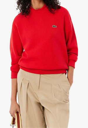 Personne portant un pull rouge Lacoste avec un pantalon plissé beige, tenant dans une main une bandoulière de sac à main beige et marron rayée.